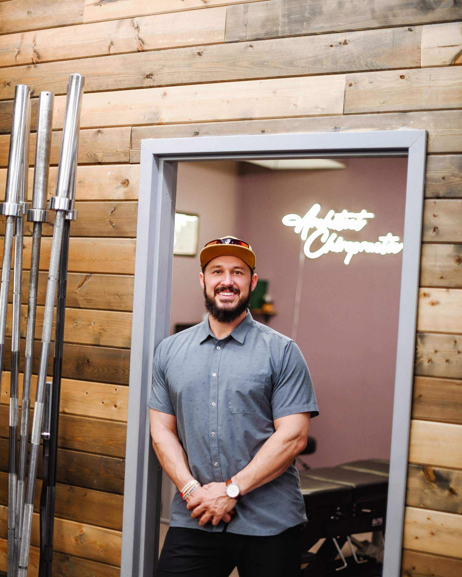 Brent standing in a doorway, hands clasped, smiling, with barbells racked on a wood plank wall beside him.
