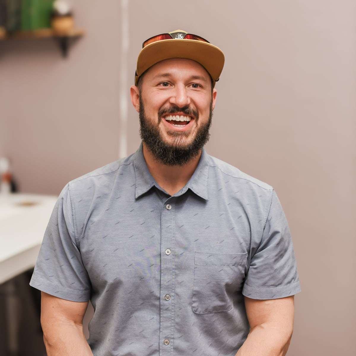 Brent laughing in front of a mauve wall, in a gray-blue button shirt and a tan cap.