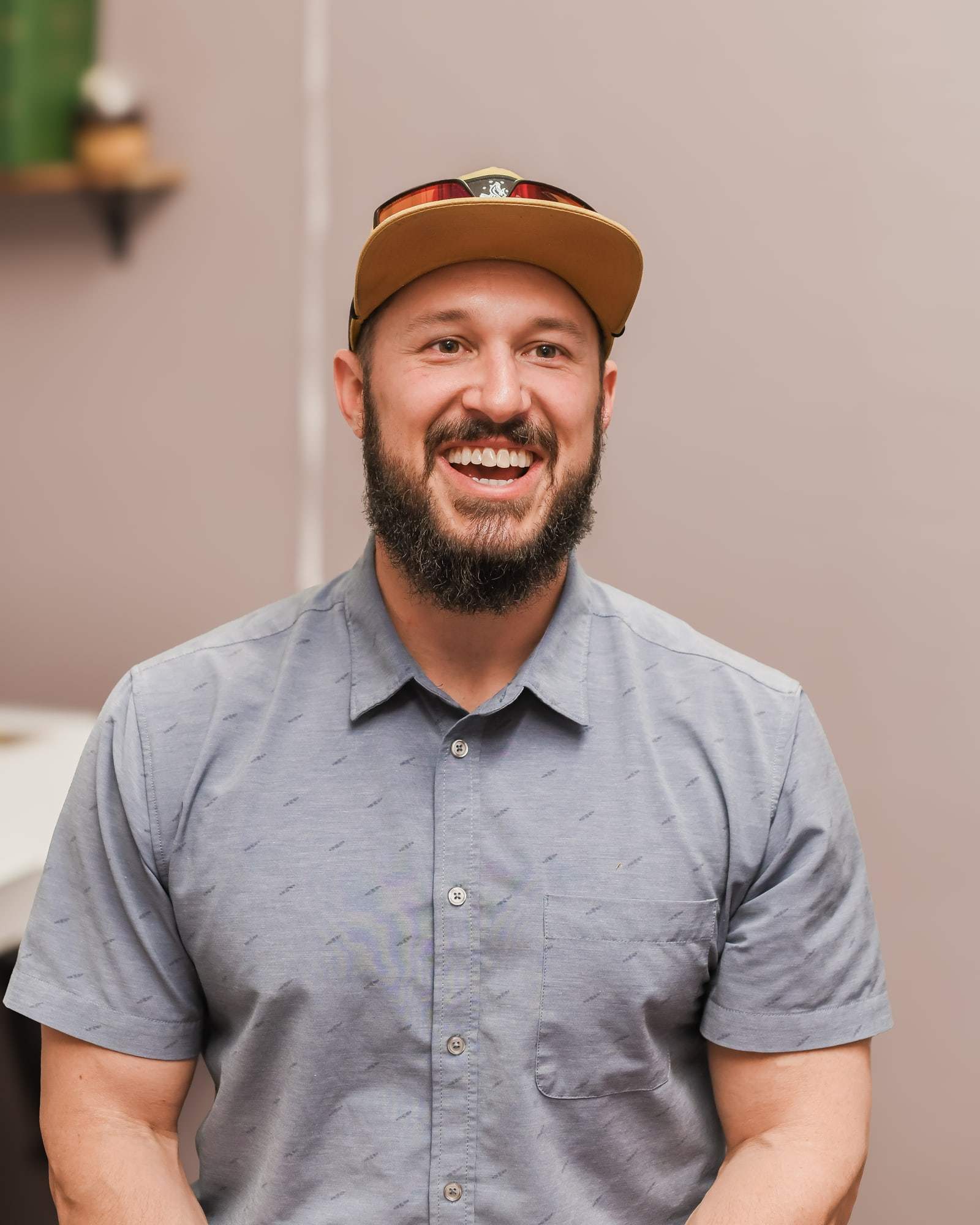 Dr. Brent Jenogan laughing, in a gray-blue button shirt and a tan cap.